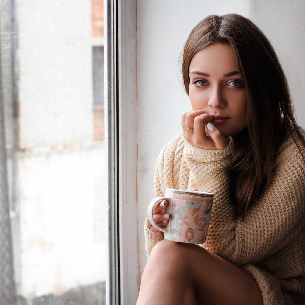Молодая девушка в свитере сидит с чашкой на подоконнике