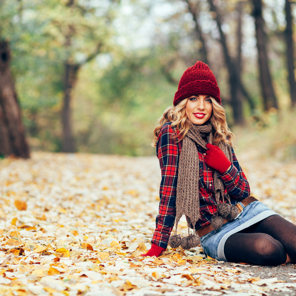 Молодая улыбающаяся девушка в красной шапке сидит на листве осенью