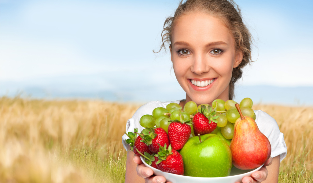 Smiling young lady with a plate with apples, grapes, strawberries and a pear in hands