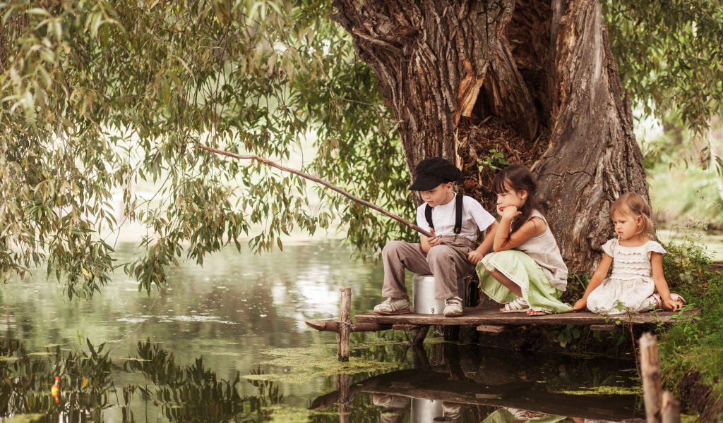 Две маленькие девочки и мальчик сидят на берегу пруда с удочкой