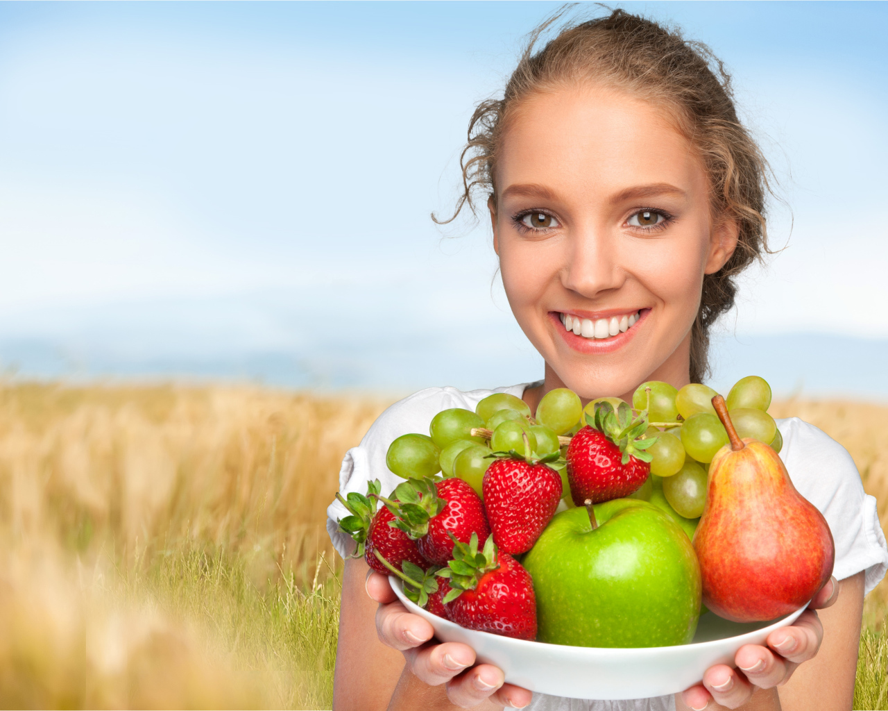 Улыбающаяся девушка шатенка с тарелкой с яблоками, виноградом, клубникой и грушей в руках