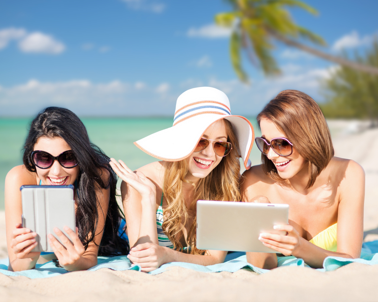 Три улыбающиеся девушки загорают на пляже с планшетами в руках