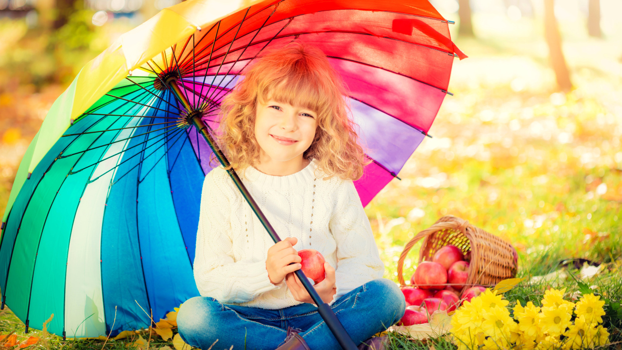 Улыбающаяся девочка с корзиной яблок сидит под разноцветным зонтом