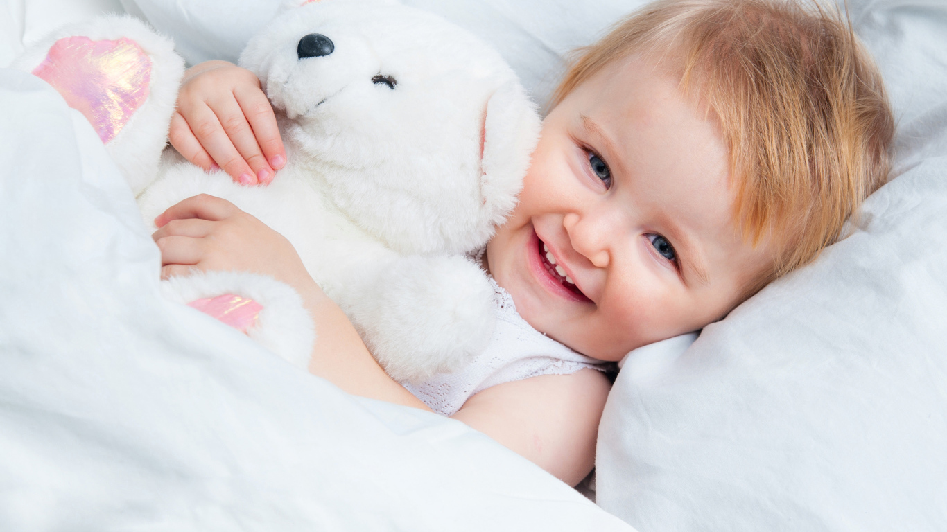 Улыбающийся ребенок лежит в кровати с белым плюшевым медвежонком