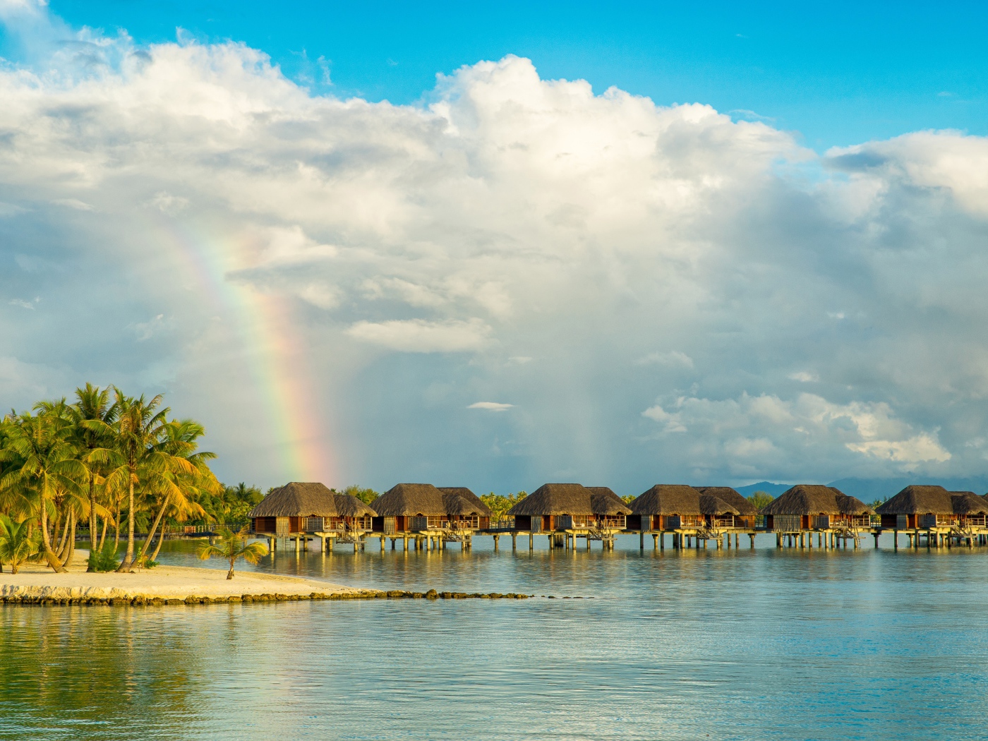 Bungalow in the water on a tropical beach under a cloudy sky with a rainbow
