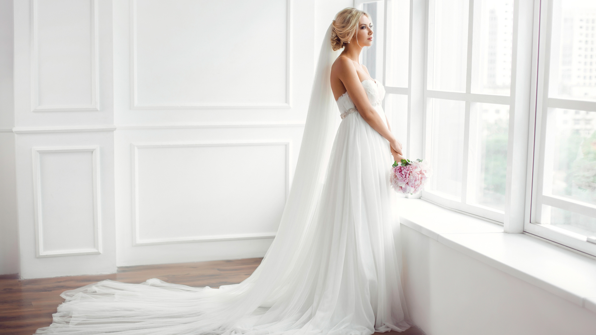 Красивая девушка в белом свадебном платье с букетом стоит у окна
