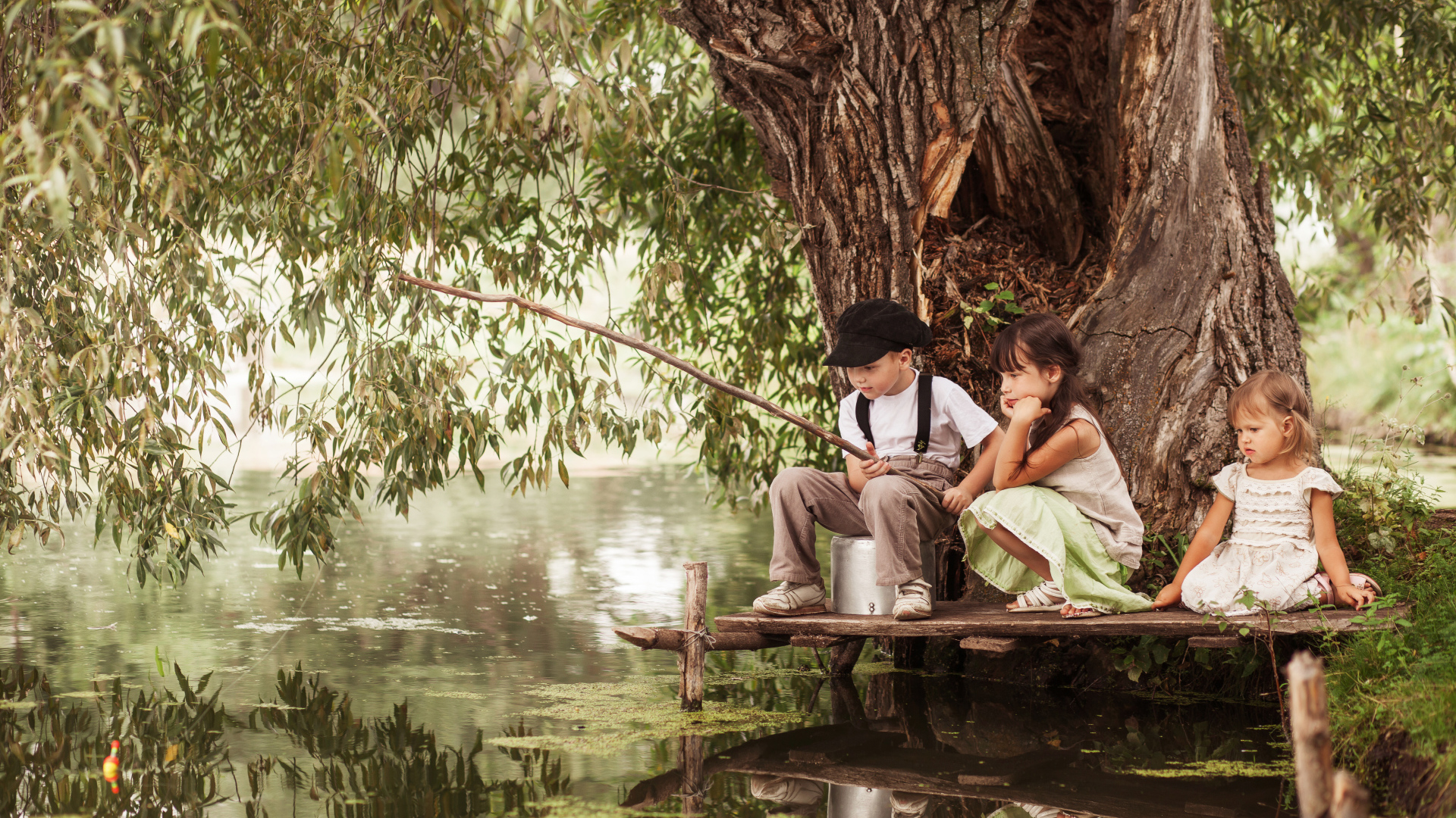 Две маленькие девочки и мальчик сидят на берегу пруда с удочкой