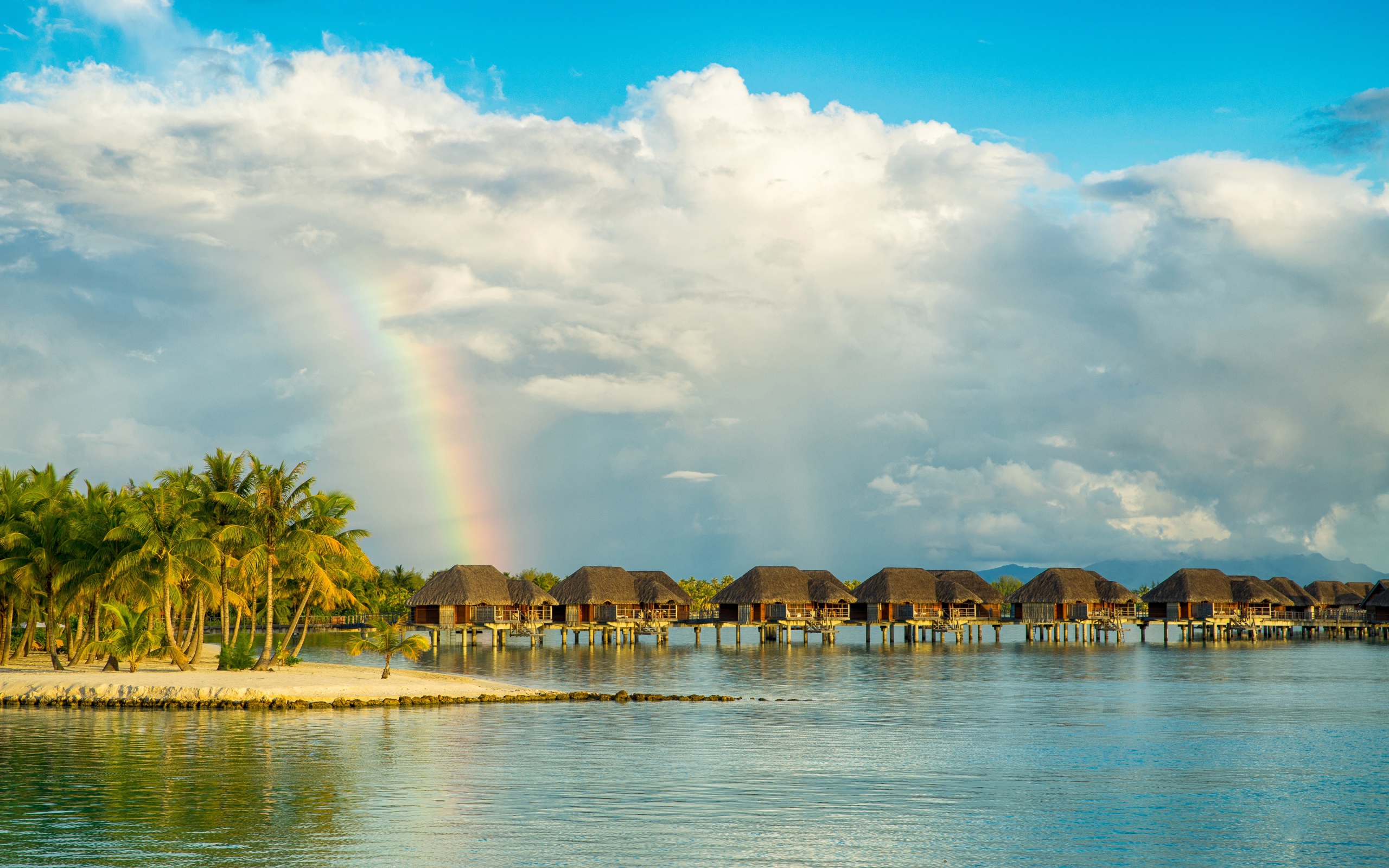 Бунгало в воде на тропическом пляже под облачным небом с радугой