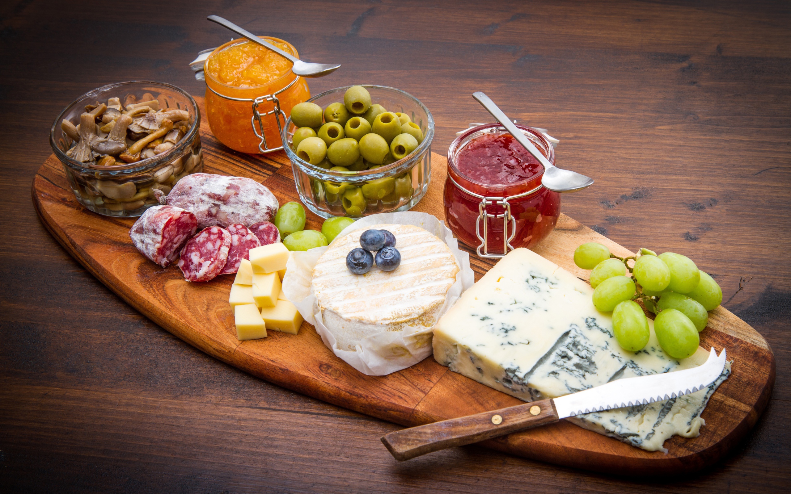 Сыр, грибы, маслины, колбаса, виноград и джем на разделочной доске 