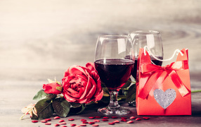 Two glasses of red wine on a table with a rose and a gift
