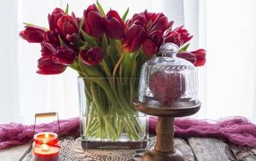 Букет красных тюльпанов в стеклянной вазе на столе со свечой и сердцем 