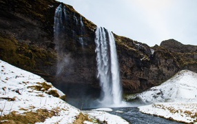 Waterfall flows down from a cliff into a river with snow-covered banks