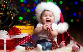 Улыбающийся малыш в новогодней шапке с подарками у елки