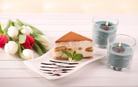 A piece of tiramisu on a table with candles and a bouquet of tulips