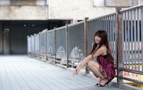 Asian girl in a sundress sits at the iron fence