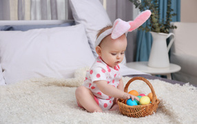 Little baby with a basket of Easter eggs is sitting on the bed.