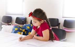 Маленькая девочка в красном платье рисует за столом