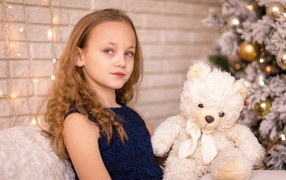 Little girl with a teddy bear sitting by a dressed up fir tree