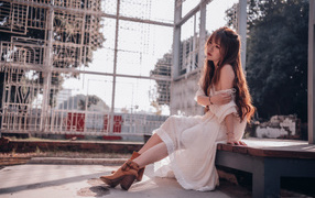 Красивая девушка азиатка в белом платье сидит на лавочке