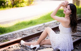 Красивая девушка азиатка в белом платье сидит на рельсах 