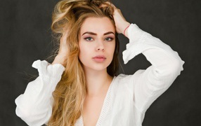 Блондинка в белой кофте с руками в голове на сером фоне 