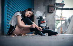 Молодая азиатка в черных чулках сидит у стены 