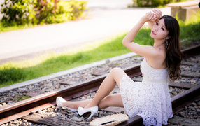 Молодая азиатка в белом платье сидит на рельсах 