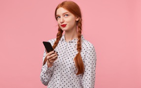 Молодая рыжеволосая девушка со смартфоном в руке на розовом фоне