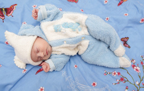 Грудной ребенок в голубом костюме лежит на кровати 