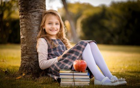 Little girl with books and an apple sits under a tree