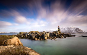 The lighthouse stands on a stone shore against the backdrop of mountains