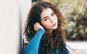 Brown-eyed girl with curly hair stands against the wall