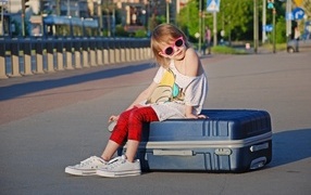 A girl in sunglasses sits on a suitcase on the road