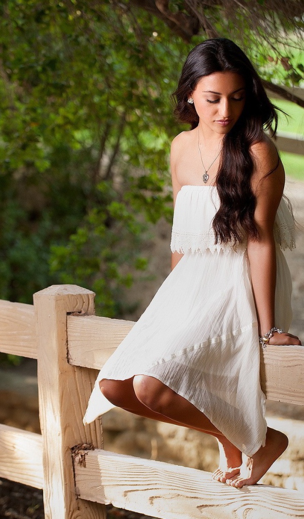 Брюнетка в белом платье сидит на заборе
