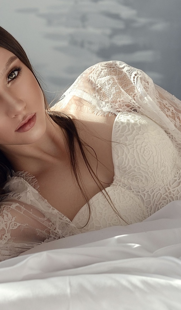 Красивая девушка брюнетка лежит на кровати в белом платье