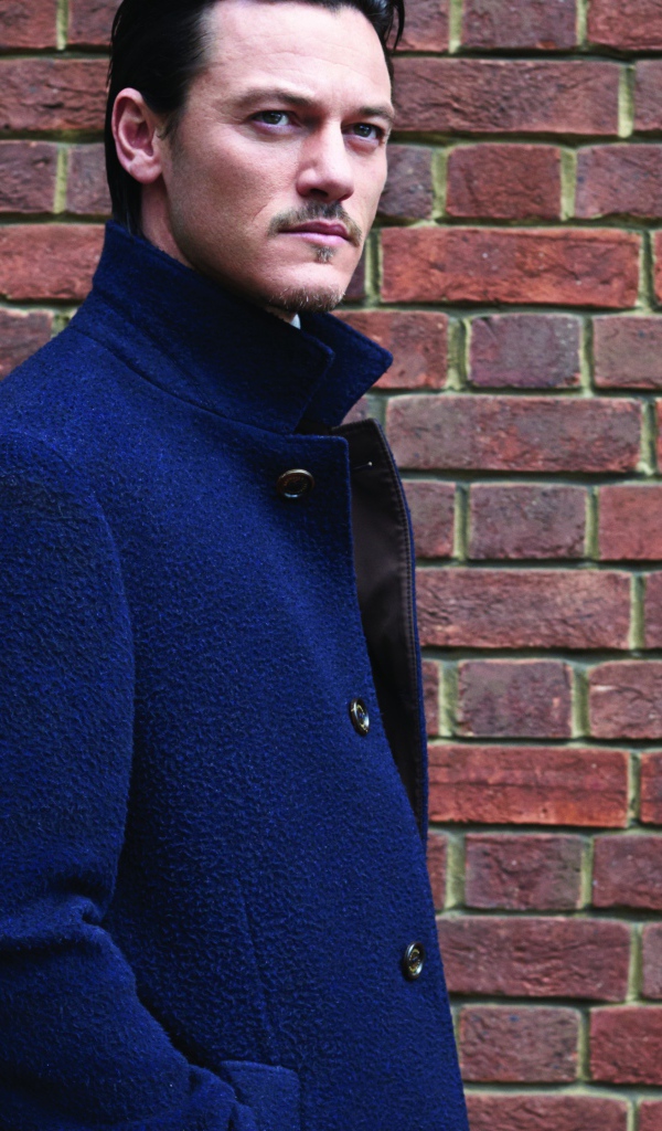 Актер Люк Эванс в синем плаще у кирпичной стены