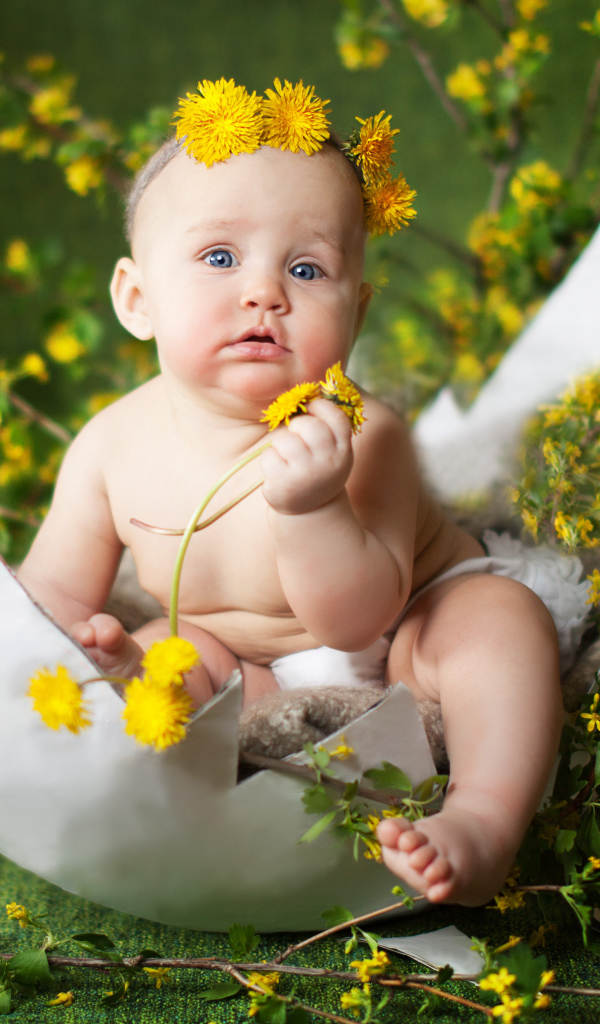 Маленький грудной ребеной сидит в яичной скорлупе с одуванчиками и ветками смородины