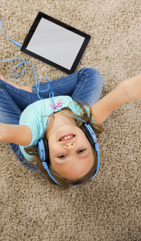 Маленькая девочка слушает музыку в наушниках сидя на ковре