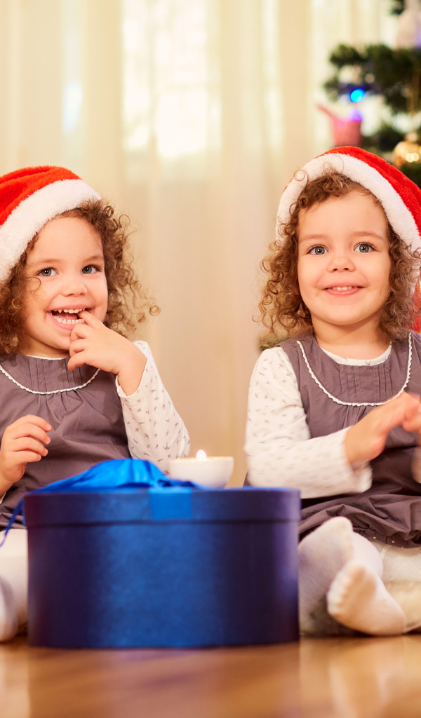 Две улыбающиеся девочки близняшки с новогодним подарком под елкой