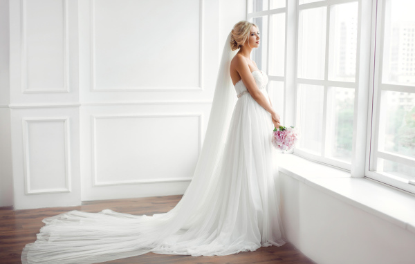 Красивая девушка в белом свадебном платье с букетом стоит у окна