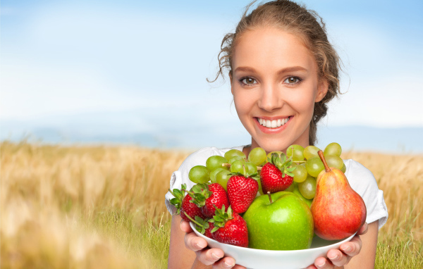 Улыбающаяся девушка шатенка с тарелкой с яблоками, виноградом, клубникой и грушей в руках
