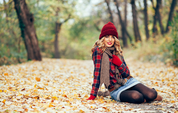 Молодая улыбающаяся девушка в красной шапке сидит на листве осенью