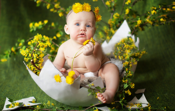Маленький грудной ребеной сидит в яичной скорлупе с одуванчиками и ветками смородины