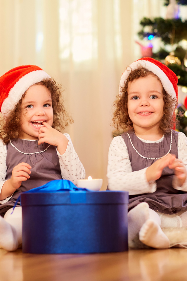 Две улыбающиеся девочки близняшки с новогодним подарком под елкой