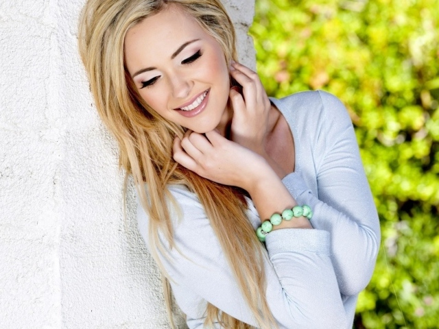 Улыбающаяся блондинка прислонилась к белой стене