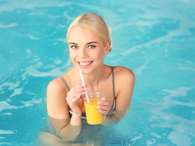 Улыбающаяся девушка блондинка в бассейне с стаканом апельсинового сока