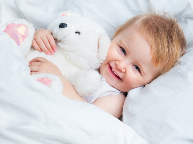 Улыбающийся ребенок лежит в кровати с белым плюшевым медвежонком
