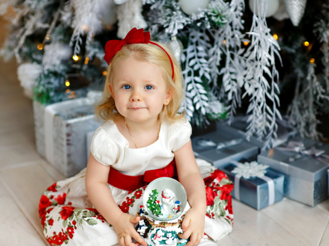Маленькая девочка блондинка сидит с подарками под новогодней елкой