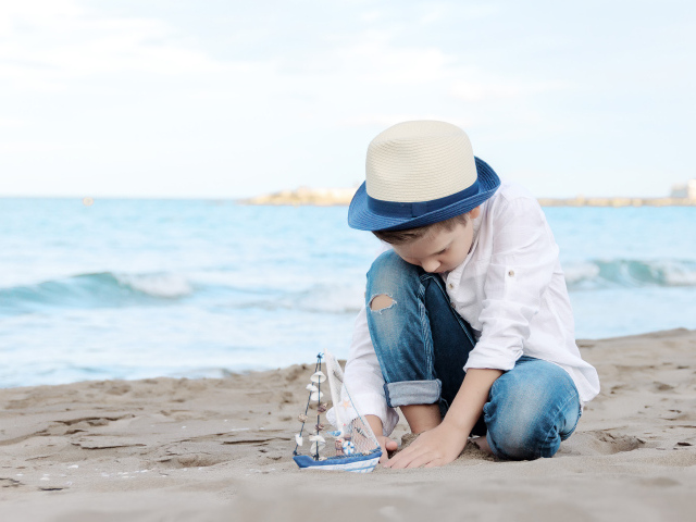 Маленький мальчик в шляпе играет с игрушечным корабликом на песке