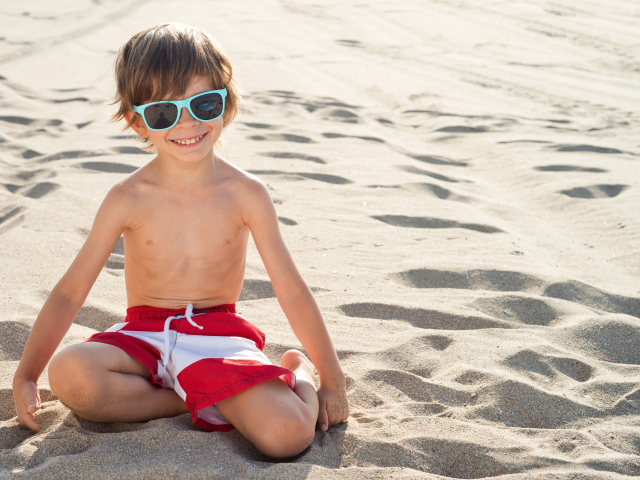 Маленький улыбающийся мальчик в очках сидит на песке